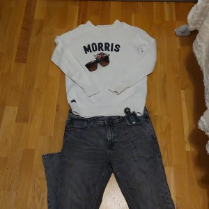 Vit stickad tröja Morris M - Vit stickad tröja från Morris med mörkblå text och brittisk flagga broderad framtill. Rund hals och ribbade muddar vid ärmslut och nederkant. Klassisk och stilren design, perfekt för dig som gillar preppy vibes.