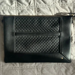 Svart kuvertväska från Edblad - Stilren svart kuvertväska från Edblad med snyggt fjällmönstrad framsida och detaljer i slätt skinn. Väskan har guldfärgad dragkedja och insida i svart textil. Perfekt storlek för att bära det viktigaste och passar till många olika looks.