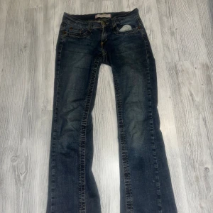Blå bootcut jeans från Vero Moda - Snygga blå jeans från Vero Moda med bootcut passform och klassisk femficksdesign. Jeansen har gul kontrastsöm och coola hjärtbroderier på bakfickorna. Materialet är jeans med slitningar och en liten reva vid fickan framtill. Perfekta för dig som gillar detaljer!