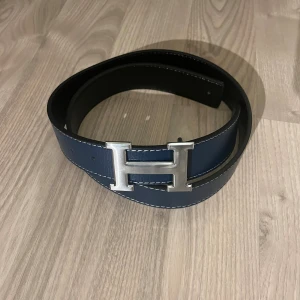 H belte blått skärp med H-spänne - Snyggt blått skärp från H belte  med ikoniskt H-spänne i silverfärgad metall. Bältet har vita kontrastsömmar och är tillverkat i exklusivt skinn. Insidan är svart, vilket ger en stilren och lyxig känsla. Perfekt accessoar för att lyfta din outfit.
