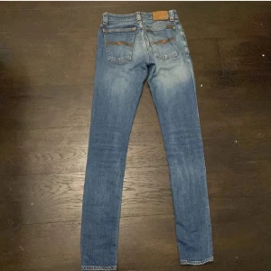 Blåa jeans från Nudie storlek W26 L30 - Snygga blå jeans från Nudie Jeans med klassisk orange söm och ikonisk bakficka. Modellen har raka ben och normal passform, tillverkade i slitstarkt bomullsjeans. Perfekta för dig som gillar en tidlös och clean look.