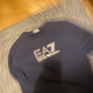 Mörkblå EA7 Emporio Armani tröja - Säljer en mörkblå tröja från EA7 Emporio Armani med stort logotryck i vitt och guld på bröstet. Klassisk rund halsringning och lång ärm. Perfekt för dig som gillar stilrena och exklusiva plagg med tydlig branding.