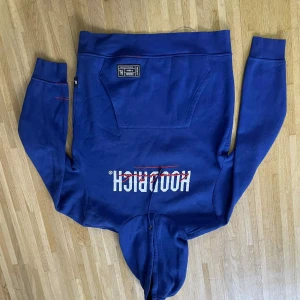 Blå HOODRICH hoodie med tryck - Snygg blå hoodie från HOODRICH med vit och röd logga framtill. Tröjan har huva med snörning, magficka och långa ärmar. Detaljer i rött på ärmen och etikett på fickan. Perfekt för dig som gillar streetwear och vill sticka ut.