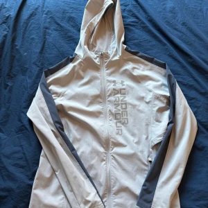 Grå vindjacka från Under Armour - Snygg grå vindjacka från Under Armour med huva och hel dragkedja. Jackan har mörkgrå detaljer längs ärmarna och axlarna samt stort logotryck på bröstet. Perfekt för träning eller när du vill ha en sportig look. Lätt och smidig modell.