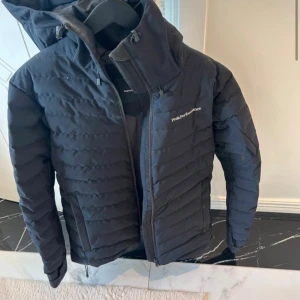 Svart dunjacka från Peak Performance - Snygg svart dunjacka från Peak Performance med huva och dragkedja framtill. Jackan har diskret logga på bröstet, quiltad design och praktiska fickor. Perfekt för kyliga dagar och enkel att matcha med olika outfits.