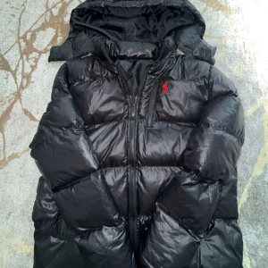 Svart pufferjacka från Ralph Lauren - Säljer en svart pufferjacka från Ralph Lauren med huva och dragkedja. Jackan har en röd broderad logga på bröstet och är riktigt fluffig och varm. Perfekt för kalla dagar och har en snygg, glansig finish. Klassisk och stilren design som funkar till det mesta.