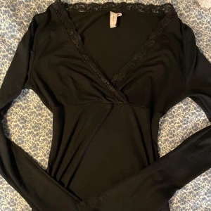 Svart omlottopp med spets från Nelly - Svart långärmad omlottopp från Nelly i storlek XS. Toppen har v-ringning med fina spetsdetaljer längs kanten och är figurnära i modellen 💕