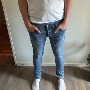 Blå skinny jeans med slitningar - Snygga ljusblå jeans med skinny passform och slitna detaljer vid benslut och fickor. Jeansen har klassisk femficksdesign, orangea sömmar och är tillverkade i jeansmaterial. Perfekt för dig som gillar en smal siluett och lite ruffig look.