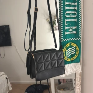 Svart quiltad axelväska med guldspänne - Snygg svart axelväska med justerbar rem och guldiga detaljer. Väskan har ett quiltat mönster med geometriska rutor på framsidan och en stilren, rektangulär form. Materialet är skinnimitation som ger en lyxig känsla. Perfekt för dig som vill ha en trendig och praktisk väska.