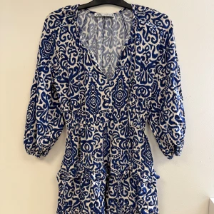 Blåvit mönstrad klänning från Zara - Supersnygg blåvit kortklänning från Zara med unikt mönster och v-ringning. Klänningen har trekvartsärm och markerad midja, perfekt för dig som gillar statement-plagg. Lätt och luftig känsla, passar perfekt till varmare dagar.