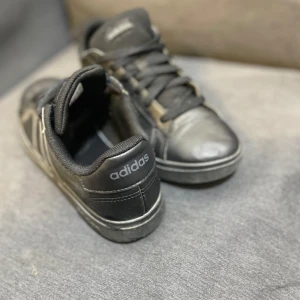 Svarta sneakers från Adidas, strl 37 - Säljer ett par svarta Adidas sneakers i storlek 37 med klassiska tre ränder på sidan och Adidas-logga på hälen. Skorna har snörning, platt sula och är tillverkade i skinn med textilfoder. Perfekt för dig som gillar stilrena och sportiga skor.