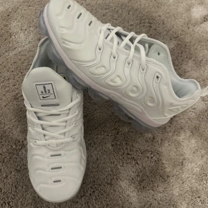 Nike Air Vapormax Plus vita sneakers - Nike Air Vapormax Plus sneakers i helvit färg med transparent sula. Snygg, futuristisk design med vågiga detaljer på ovandelen och klassisk snörning. Märkeslogga på plösen och hälen. Ovandel i syntetmaterial och mesh för skön komfort. Helt nya 