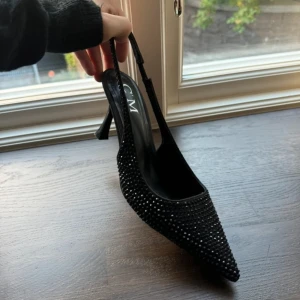 Svarta glittriga pumps från CM Paris - Svarta pumps med spetsig tå från CM Paris, täckta med små svarta glittriga stenar. Skorna har en låg klack och öppen häl med smal rem. Perfekt för dig som gillar att sticka ut med eleganta detaljer och glam.