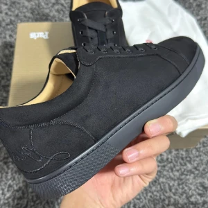 Svarta sneakers från Christian Louboutin - Snygga svarta sneakers från Christian Louboutin med diskret broderad logga på hälen. Skorna har en rund tå, platt sula och är tillverkade i mjuk mocka med svarta snören. Insidan är beige och de har metallöglor på sidan för ventilation. Perfekt för dig som gillar stilrena och lyxiga detaljer.