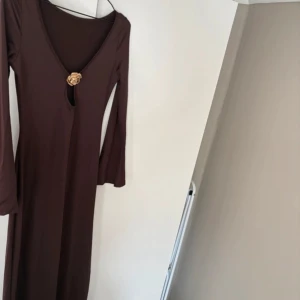 Brun långklänning - Säljer en elegant brun långklänning med v-ringning och långärmade ärmar. Klänningen har en snygg blomformad detalj framtill vid halsen som ger en unik touch. Perfekt för dig som gillar stilrena och feminina plagg.