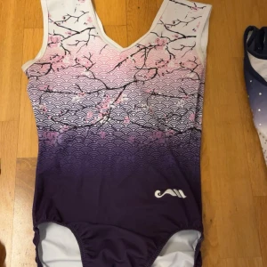Lila och vit gymnastikdräkt med körsbärsblommor - Supersnygg gymnastikdräkt från Milano med lila och vit färgskiftning, dekorerad med japanskinspirerat vågmönster och körsbärsblommor i rosa. V-ringad hals och bekväm passform, perfekt för träning och tävling