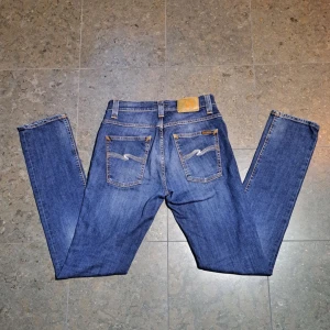 Nudie Jeans  - Säljer ett par klassiska blå jeans från Nudie Jeans med snygga kontrastsömmar och broderade bakfickor. Modellen har normal passform och raka ben, tillverkade i bomull. Perfekta för dig som gillar stilrena och tidlösa jeans. Priset kan diskuteras!