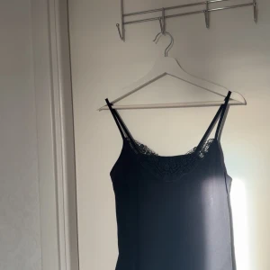 Svart linnetopp med spets - Snygg svart linnetopp med tunna axelband och dekorativ spets längs halsringningen. Toppen har en enkel och stilren design som passar perfekt till jeans eller kjol. Materialet känns mjukt och lätt mot huden, och spetsen ger en extra touch av elegans.