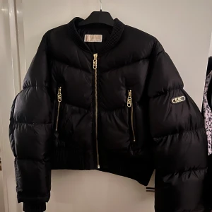 Svart pufferjacka från Michael Kors - Säljer en svart pufferjacka från Michael Kors med guldiga MK-detaljer på dragkedjor och ärmen. Jackan har ribbad krage och muddar, samt flera fickor med snygga dragkedjor. Perfekt för dig som gillar en oversized look och vill sticka ut med exklusiva detaljer.