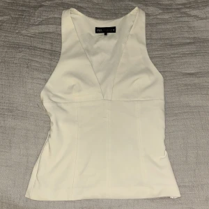 Vit v-ringad linnetopp från Zara - Säljer en stilren vit linnetopp från Zara i storlek S. Toppen har en djup v-ringning och breda axelband, vilket ger en snygg och modern siluett. Materialet är mjukt och har en lätt glans. Perfekt att matcha med jeans eller kjol för en fräsch look.