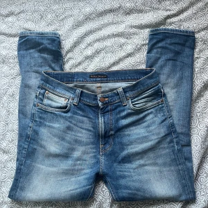 Blå skinny jeans från Nudie Jeans - Snygga blå jeans från Nudie Jeans i modellen skinny med klassisk femficksdesign och orangea sömmar. Jeansen har en ljus tvätt med slitningar och Nudie-logga på bakfickan. Perfekt passform och stilren look för dig som gillar smala jeans.  W29 L28