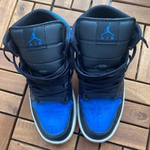 Nike Air Jordan 1 svart/blå US8 - Säljer ett par Nike Air Jordan 1 i svart och blått skinn med klassisk swoosh på sidan och blå detaljer på sulan. Höga sneakers med svart snörning och ikonisk Jumpman-logga på plösen. Perfekta för dig som vill sticka ut med en streetstyle-look.