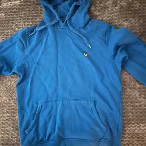 Blå hoodie från Lyle & Scott - Säljer en blå hoodie från Lyle & Scott med klassisk gul logga på bröstet. Tröjan har huva med snörning och en stor magficka framtill. Tillverkad i mjuk bomull, perfekt för chill dagar eller när du vill vara extra bekväm.