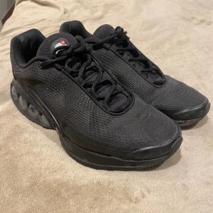 Nike Air Max Dn - Svarta Nike Air Max sneakers med mesh-överdel och synliga luftbubblor i sulan. Skorna har snörning, rund tå och en chunky sula med röda detaljer undertill. Perfekta för dig som gillar sportig och modern stil.