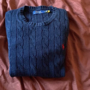 Marinblå stickad tröja Polo Ralph Lauren - Kabelstickad marinblå tröja från Polo Ralph Lauren med rund halsringning och klassisk röd broderad logga på bröstet. Tröjan har ribbade muddar och är långärmad, perfekt för lager-på-lager under kyliga dagar.