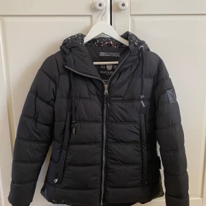 Svart pufferjacka från Navahoo storlek S - Snygg svart pufferjacka från Navahoo med quiltad design, huva med dragsko och coolt prickigt innerfoder. Jackan har dragkedja framtill, ribbade muddar och praktiska fickor. Perfekt för kalla dagar och ger en modern look. Fint skick, endast använd en  säsong.. Nypris 1200kr.