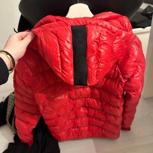 Röd dunjacka från Peak Performance - Säljer en snygg röd dunjacka från Peak Performance. Jackan har en dragkedja framtill och en huva för extra värme. Perfekt för kyliga dagar med sin stilrena design och praktiska fickor. Märkesdetaljer på ärmen ger en sportig touch.