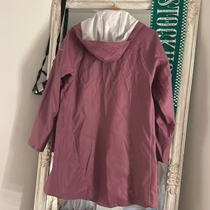 Rosa regnjacka från Cross Sportswear - Lång, rosa regnjacka från Cross Sportswear med huva och vita detaljer. Jackan har dragkedja, tryckknappar och två stora fickor framtill. Perfekt för regniga dagar och har en clean, stilren look.