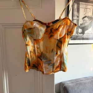 Färgglad korsett från PrettyLittleThing - Säljer en snygg korsett-topp från PrettyLittleThing med tunna axelband. Toppen har ett färgstarkt mönster i orange, gul, blå och beige toner och är sydd i ett mjukt, lätt material. 