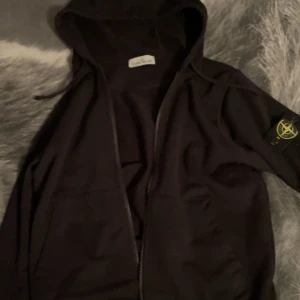 Svart zip up från Stone Island - Svart hoodiejacka från Stone Island med ikonisk patch på ärmen. Jackan har dragkedja framtill, huva med snörning och två fickor. Tillverkad i mjuk bomull för en skön och avslappnad stil. Perfekt för dig som gillar streetwear och vill ha något stilrent.
