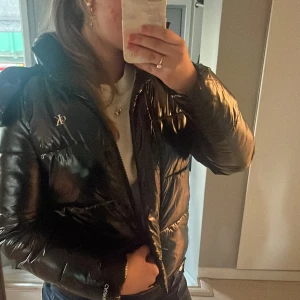 Glansig Jacka från Calvin Klein🤗 - Säljer en svart, glansig pufferjacka från Calvin Klein med huva och dragkedja. Jackan har CK-logga på bröstet och stor Calvin Klein Jeans tryck på insidan. Använd fåtal gånger!. Nypris-2200kr. Perfekt för dig som vill ha en snygg och varm jacka 💕