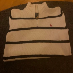 Randig half zip tröja Polo Ralph Lauren - Vit stickad tröja från Polo Ralph Lauren med svarta horisontella ränder och röd broderad logga på bröstet. Tröjan har ribbad krage, muddar och nederkant samt en half zip-dragkedja vid halsen. Tillverkad i 100% bomull.