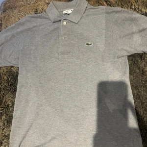 Grå pikétröja från Lacoste XS - Klassisk grå pikétröja från Lacoste i storlek XS. Saknas en knap vid kragen. Tröjan har korta ärmar, krage med två knappar och den ikoniska gröna krokodilloggan broderad på bröstet. Perfekt basic med clean look och skön passform.
