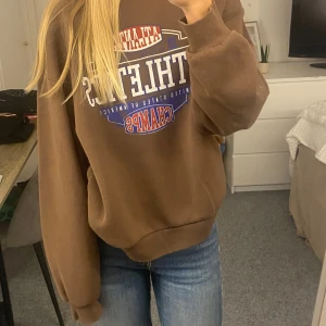 Brun sweatshirt från Gina Tricot XS - Säljer en brun sweatshirt från Gina Tricot i storlek XS med tryck i vitt, blått och rött framtill där det står 'Atlanta Athletics Champs'. Tröjan har rund hals, långa ärmar och är gjord i mjuk bomullsblandning. Perfekt för en chill och sportig look, lite oversized. 