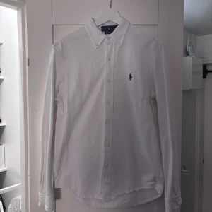 Vit Oxford skjorta, Ralph Lauren - Vit Oxford skjorta från Ralph Lauren med klassisk button-down krage och blå broderad logga på bröstet. Tillverkad i lätt mesh-bomull som känns luftig och skön. 