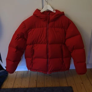 Röd pufferjacka från Lacoste L/XL - Riktigt fet röd pufferjacka som är varsamt använd från Lacoste i storlek L/XL. Jackan har huva, dragkedja och broderad Lacoste-logga både fram och stort på ryggen. Perfekt för kalla dagar när du vill sticka ut och hålla dig varm. Ny pris 4000.