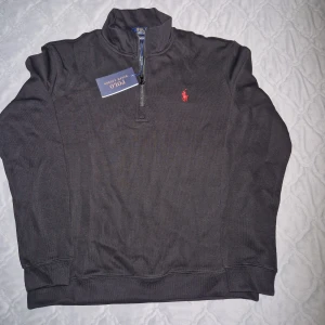 Svart half zip tröja Polo Ralph Lauren - Snygg svart långärmad tröja från Polo Ralph Lauren med röd broderad logga på bröstet. Tröjan har en half zip-dragkedja vid halsen och är tillverkad i mjuk bomull. Perfekt för lager-på-lager och chill stil.