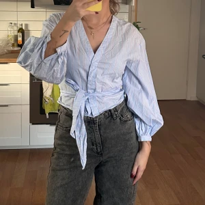Blå omlottblus med knyt och puffärm - Säljer en ljusblå omlottblus med tunna röda ränder. Blusen har v-ringning, knyt i midjan och ballongärmar med knapp vid ärmslut. 💓💓