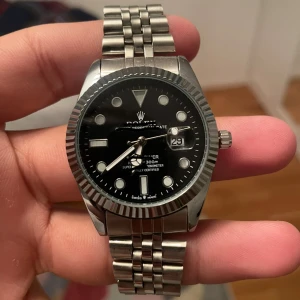 (R£P) ROL£X silver klocka män - Snygg klocka med svart urtavla och silvrig boett och länk i rostfritt stål. Klockan har tydliga index, datumfönster med förstoringsglas och klassisk räfflad bezel. Länken är robust och har ROL£X-loggan på spännet. Perfekt för dig som gillar stilrena accessoarer.