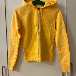 Gul hoodie med dragkedja och huva - Gul hoodie med huva och hel dragkedja framtill. Tröjan har två fickor och ribbade muddar vid ärmslut och nederkant. Tillverkad i 100% bomull, perfekt för dig som gillar färgglada och bekväma plagg. Uppskattar att storleken är 36 i europeiskt mått.