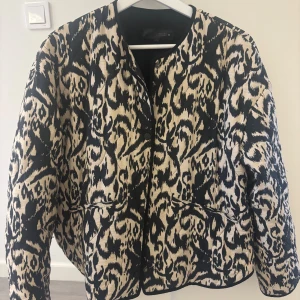 Mönstrad quiltad jacka Zara - Säljer en snygg quiltad jacka från Zara med mönster i svart och beige. Jackan har rund halsringning, knappar framtill och två fickor. Perfekt statement-plagg för dig som vill sticka ut.