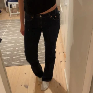 Mörkblå bootcut jeans med fickdetaljer - Säljer ett par mörkblå jeans med bootcut passform och låg midja. Jeansen har klassiska fickor fram och bak, där bakfickorna har snygga lock med silverfärgade knappar. Materialet är jeans och de sitter riktigt snyggt på kroppen.