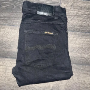 Nudie Grim Tim jeans  - Svarta jeans från Nudie Jeans med klassisk femficksdesign och diskret logga på bakfickan. Modellen är tight och sitter snyggt längs benen. modell Grim Tim. Midja 40cm, längd 100cm. JAG SKICKAR EJ FLER BILDER NÄR JAG BÄR JEANSEN! för storleksguide hänvisar jag till måtten och för bättre inblick i passformen rekommenderar jag att googla på modellnamnet :) s3 