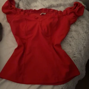 Röd offshoulder topp med volang - Säljer en snygg röd offshoulder topp med volangkant upptill. Toppen har en figurnära passform och markerad midja som ger en schysst siluett. Perfekt för dig som vill sticka ut med en färgstark look. Materialet känns mjukt och följsamt mot kroppen.