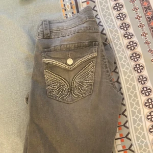 Grå jeans med broderad bakficka - Snygga grå jeans med unik broderad design på bakfickan och silverfärgad knapp. Jeansen har klassisk femficksmodell och är tillverkade i mjukt denimtyg. Perfekt för dig som gillar detaljer och vill sticka ut lite extra.