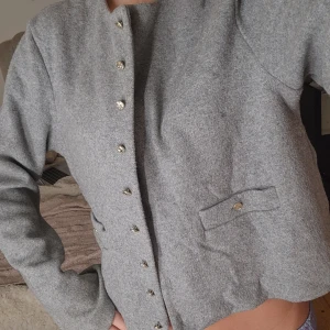 Grå ullkofta med guldknappar från H&M - Stilren grå ullkofta från H&M med rund halsringning. Jackan har dekorativa guldknappar framtill och två fickor. Perfekt för dig som gillar en clean och klassisk look med en twist.